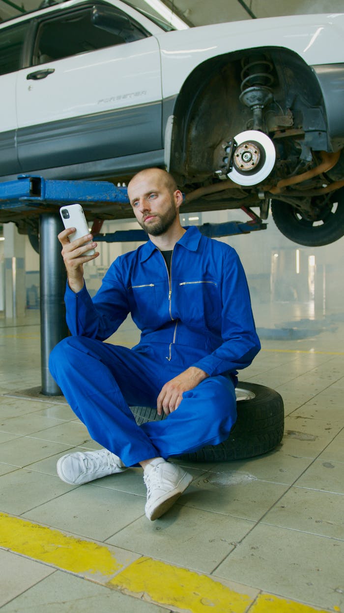 Mechanic in a blue jumpsuit sitting on a tire, using a smartphone, with a car in repair overhead.
